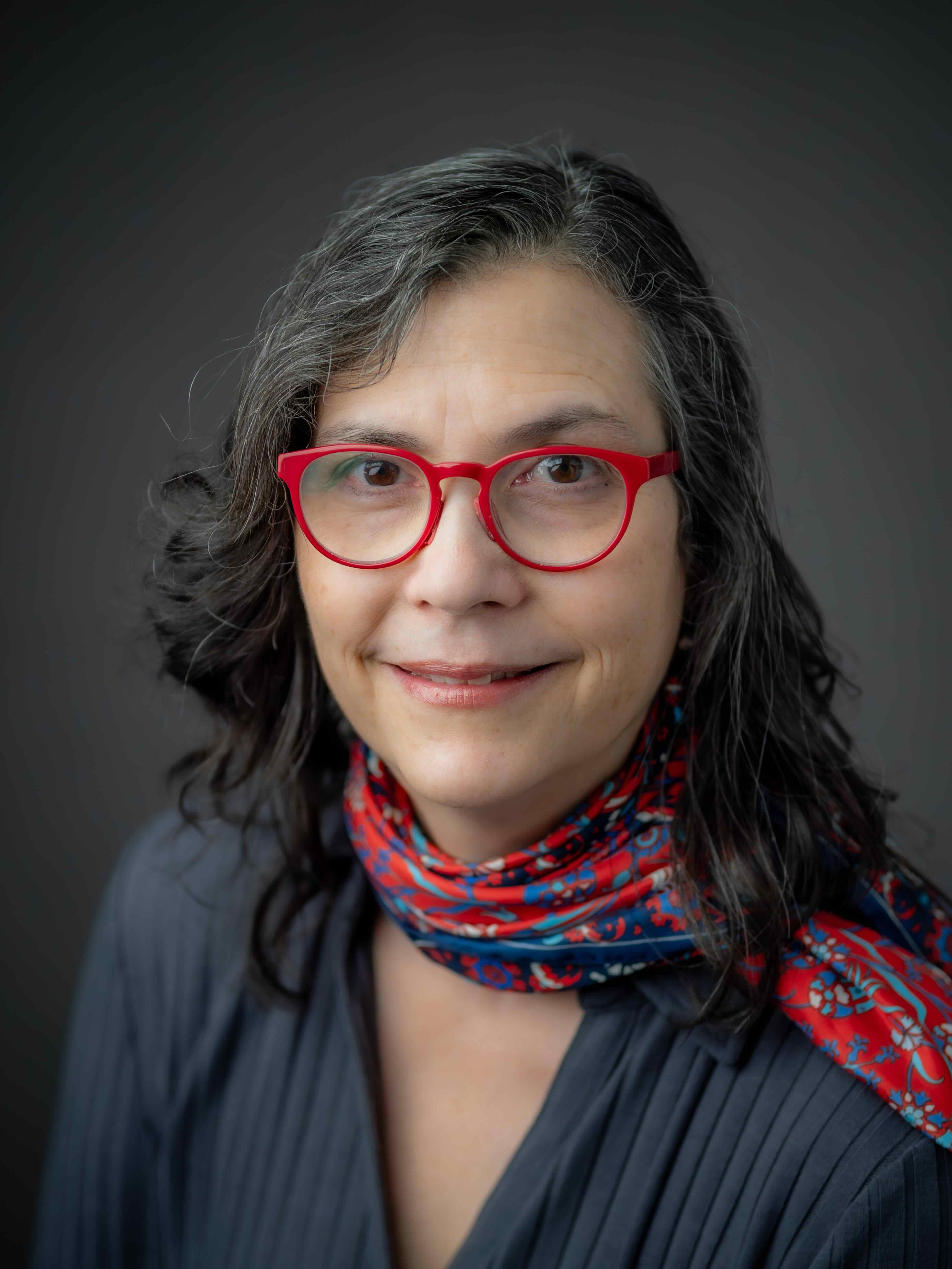 Elizabeth is has short dark hair, is smiling, wearing a white collared shirt, tortise shell glasses and silver earrings